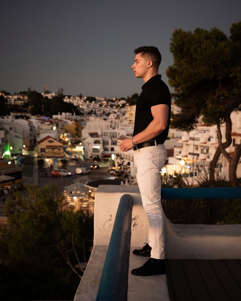 Young man in black polo and white jeans stands on a wall overlooking a Mediterranean town at dusk.