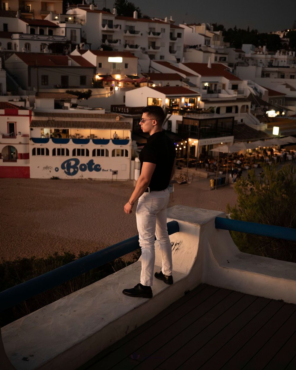 Man in black shirt and white jeans standing on a wall at night with a coastal town illuminated behind him.