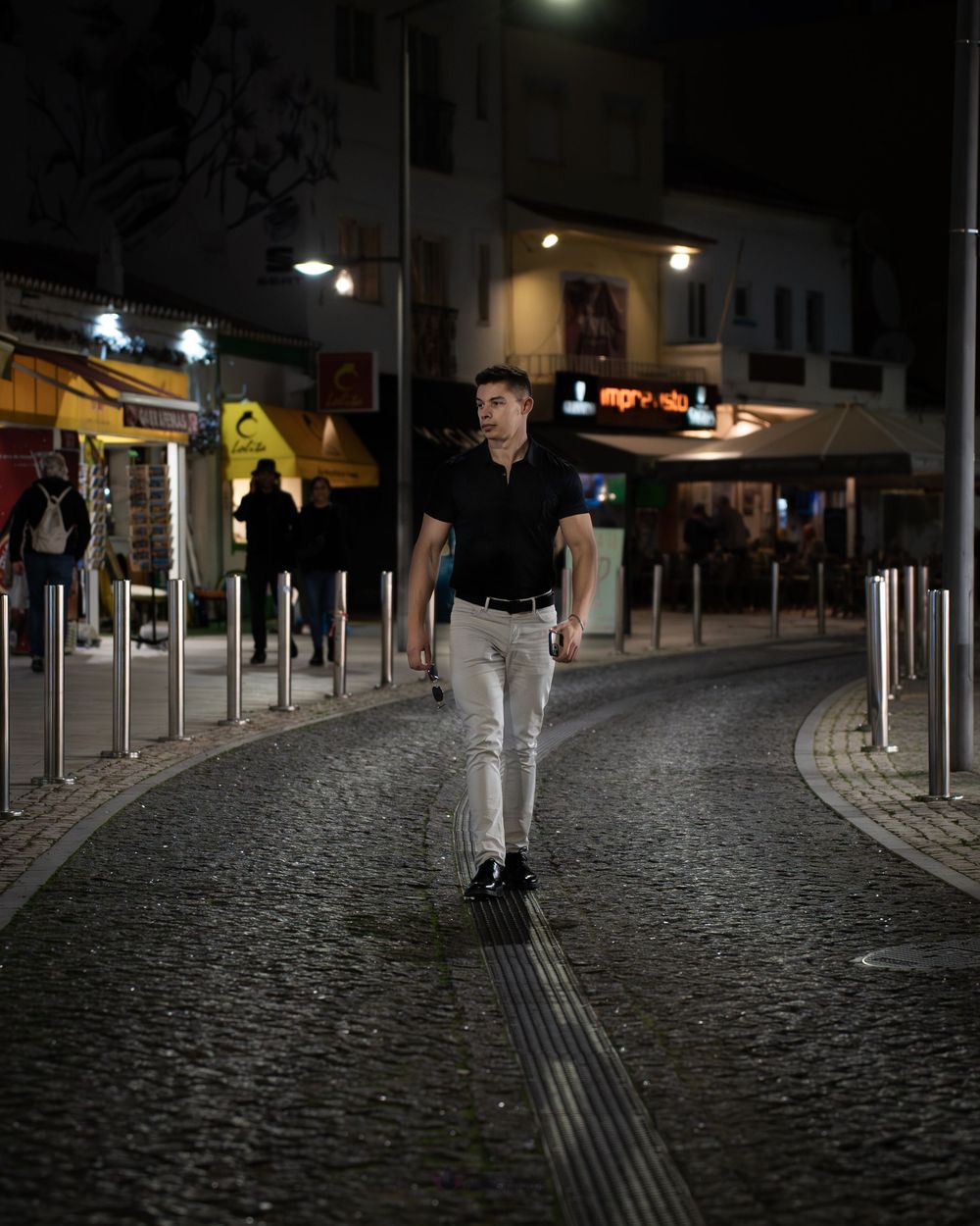 Man in black polo and white pants walks cobblestone street at night with lit shops in background.