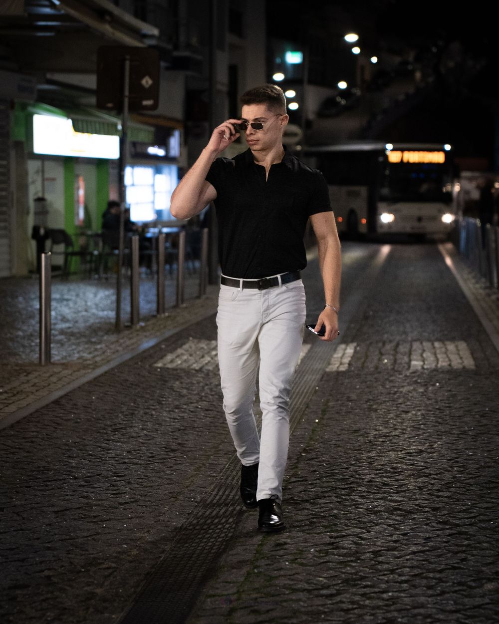 Stylish man in black polo and white jeans walking on cobblestone street at night wearing sunglasses.