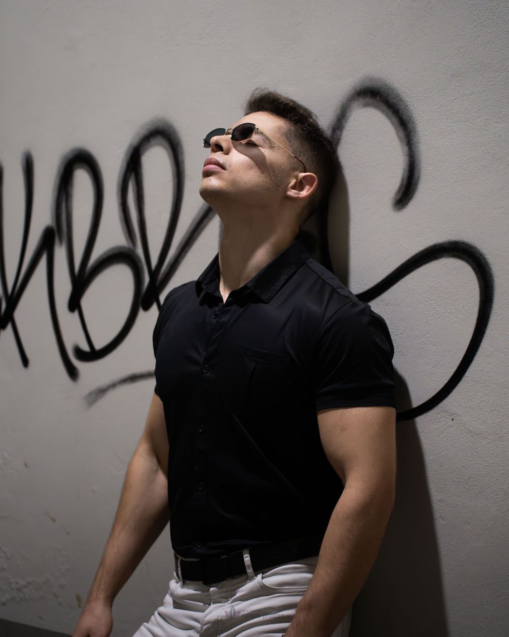 Muscular man in black polo and sunglasses leaning against graffiti wall, looking up in sunlight.