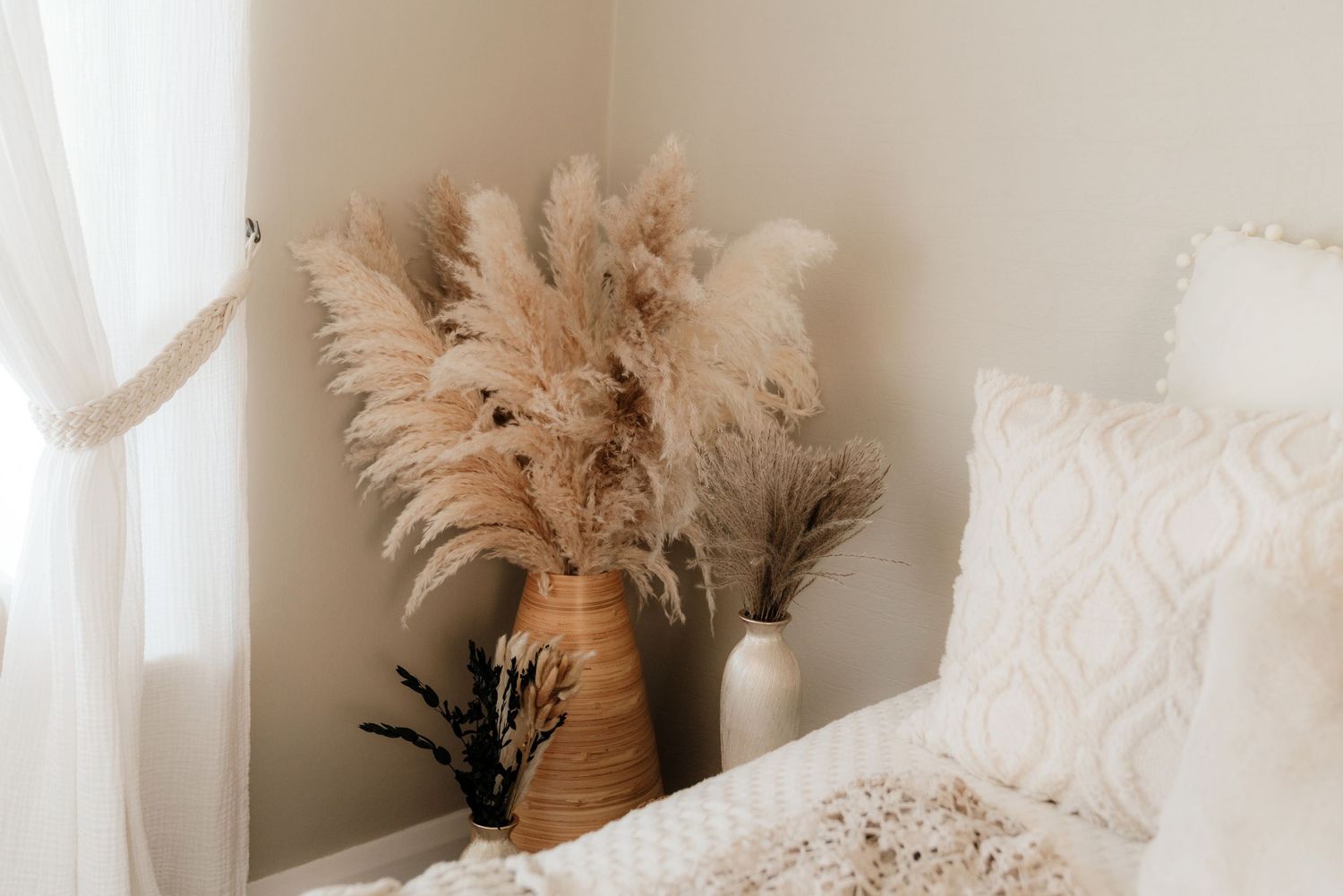 Boho bedroom corner with pampas grass in woven vase and white ceramic vase beside cozy white bedding.
