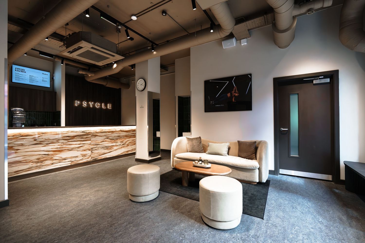 Modern Psycle fitness studio reception area featuring a marble front desk, white sofa, ottomans, and industrial ceiling details.