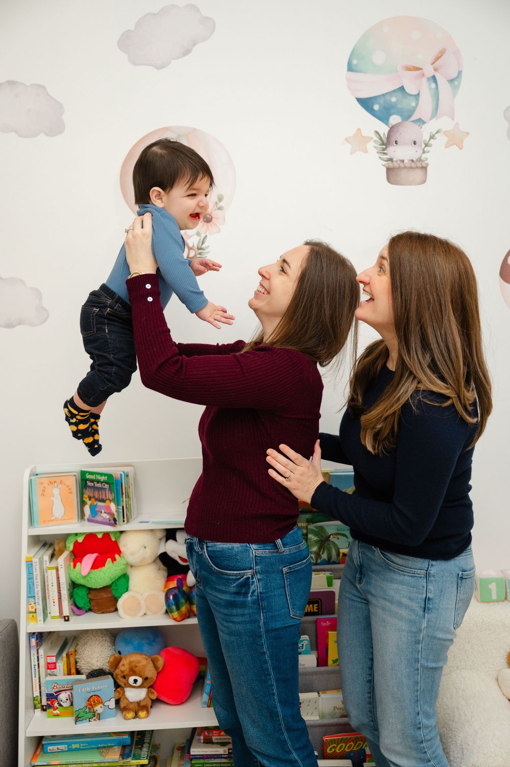 Two mothers lifting and playing with their laughing baby in a colorful nursery with wall decals.