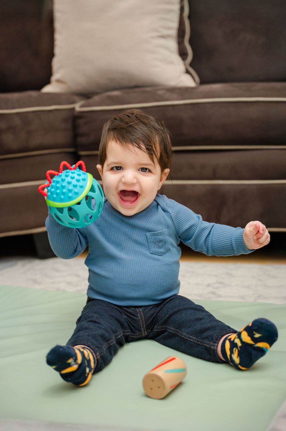 Happy toddler boy in blue shirt playing with colorful toy ball on floor near brown sofa.
