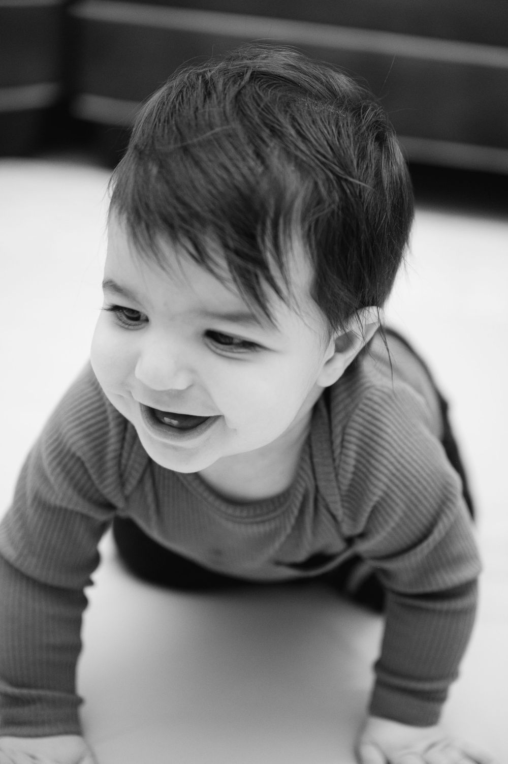 Smiling baby in gray ribbed onesie crawling on floor, black and white photo.