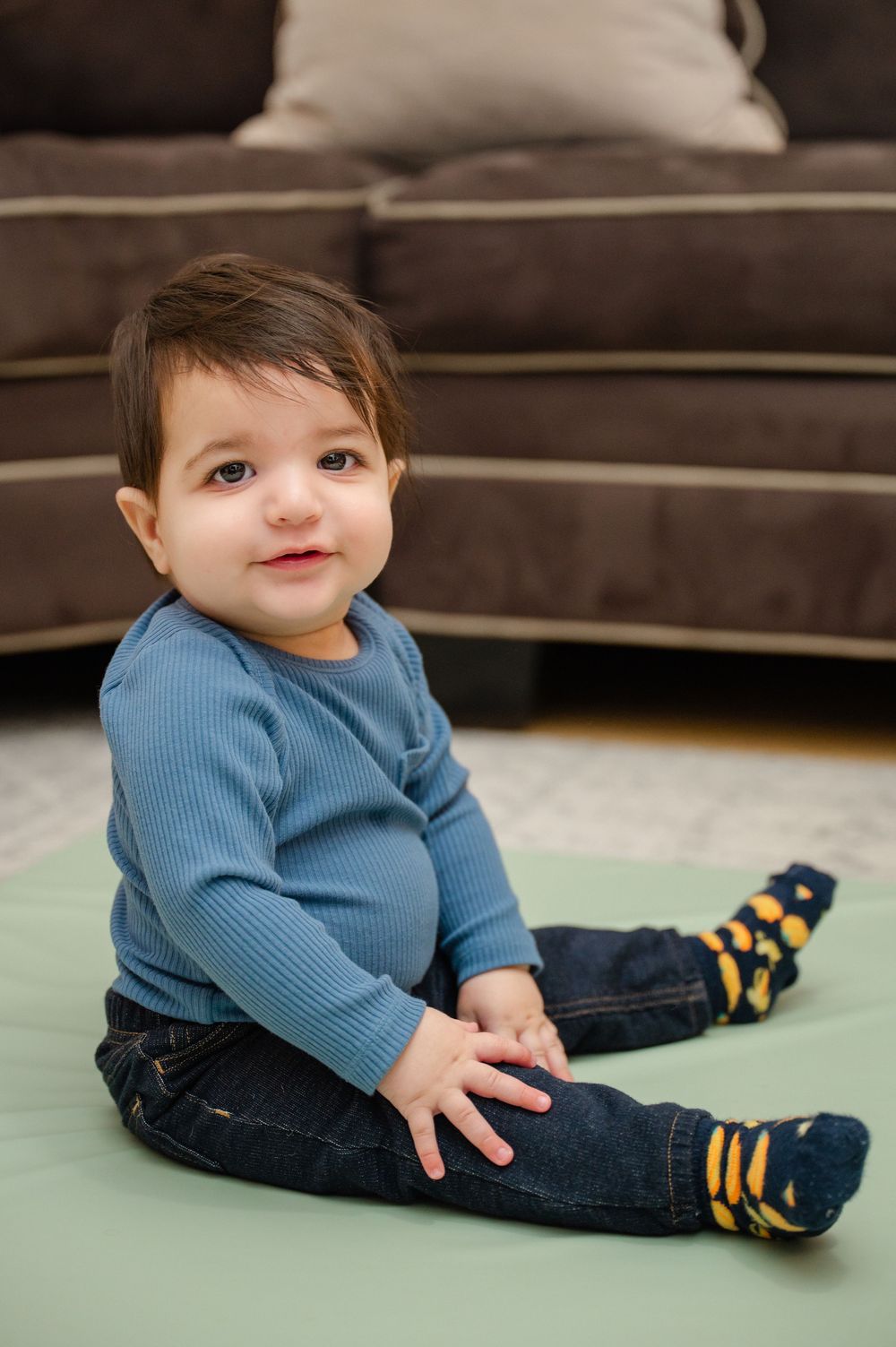 Cute baby boy sitting on green mat in blue ribbed shirt, jeans, and colorful socks.
