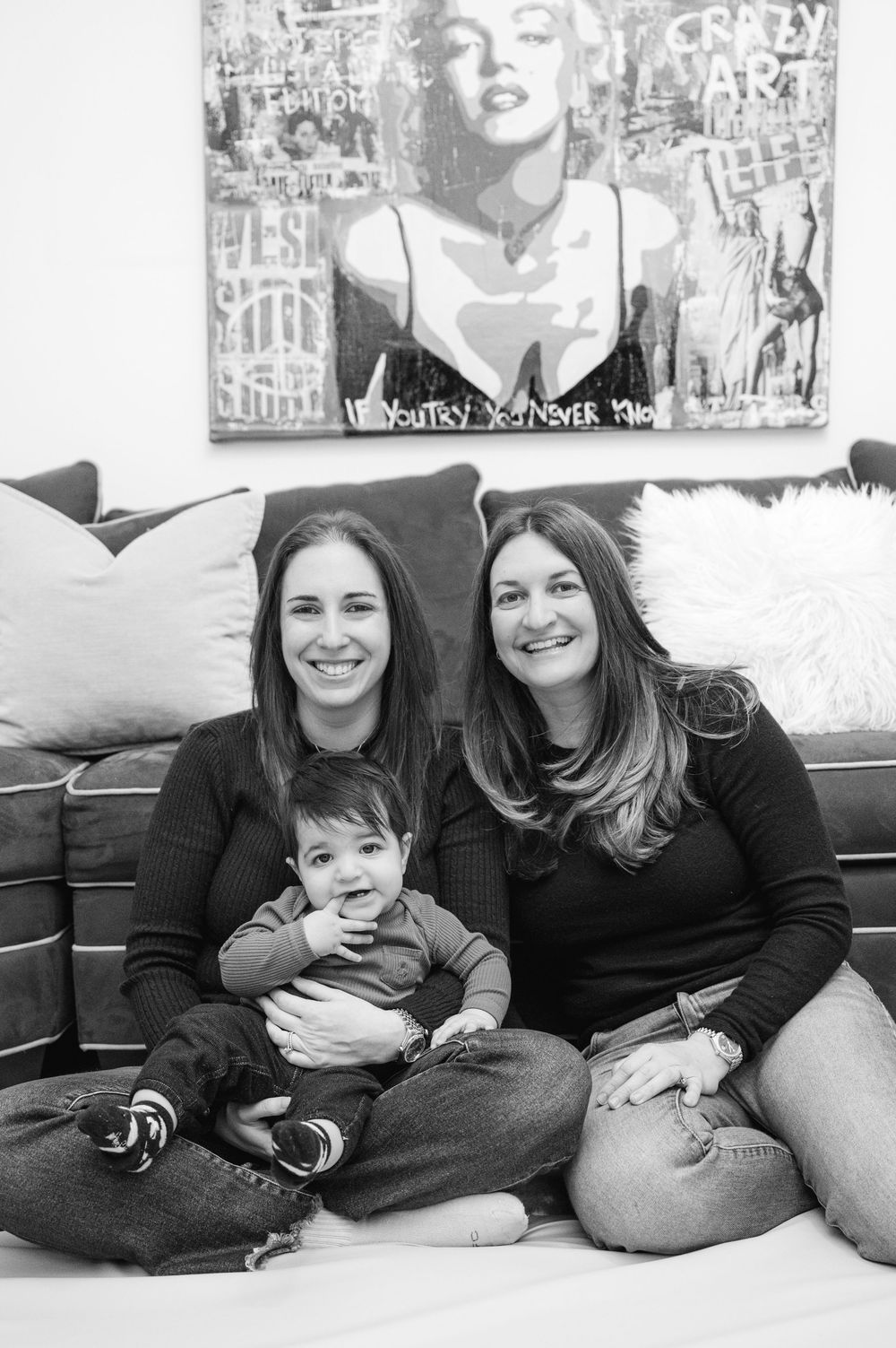 Two women smiling with an infant sitting together on a couch in a black and white photo.