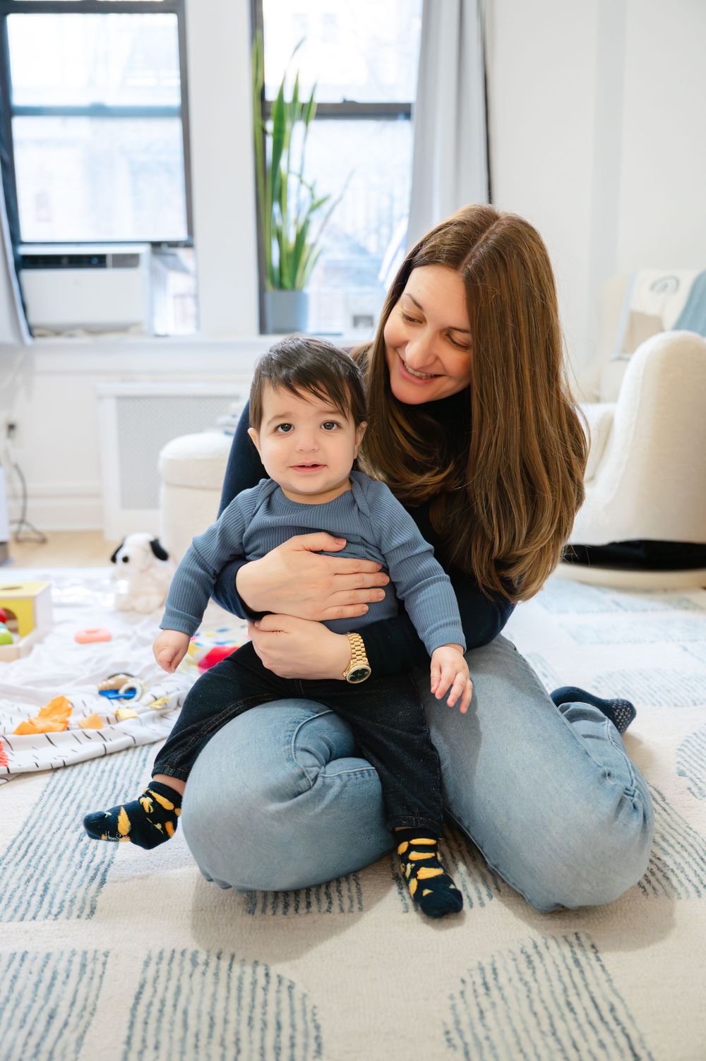 A smiling mother sits on a floor holding her baby boy in a bright, modern living room.