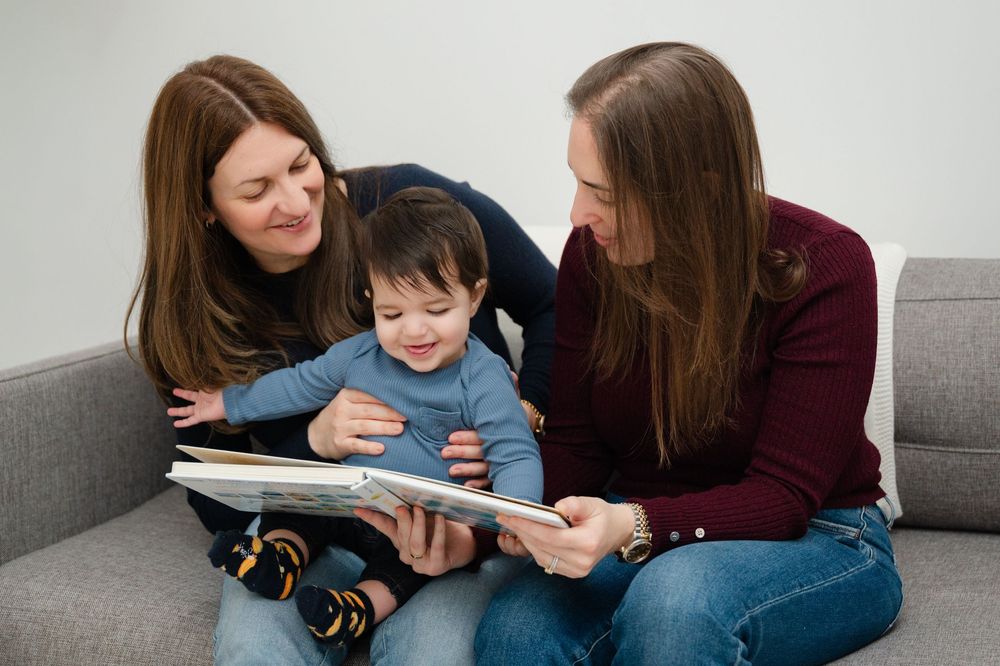 Two women sit on a gray sofa reading a picture book with a happy toddler boy.