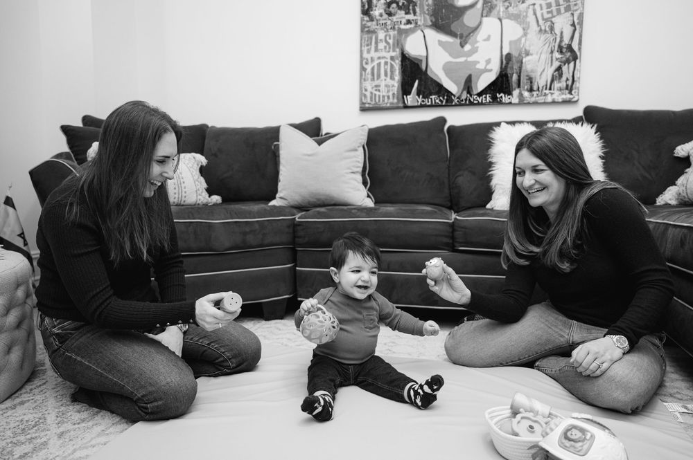 Two women sit on floor laughing with a happy baby between them near a gray couch in black and white photo.