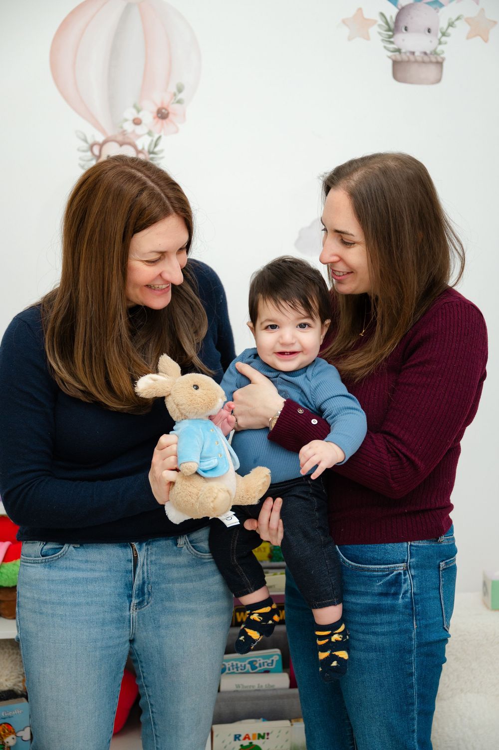 Two women smiling and holding a happy baby with a Peter Rabbit stuffed toy in a decorated nursery room.