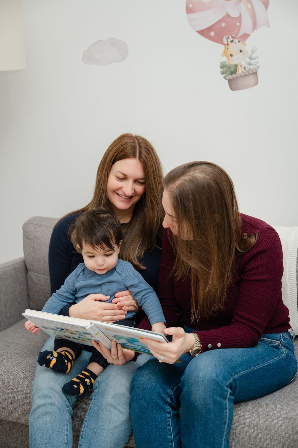 Two women sitting on a couch reading a picture book with a happy toddler in a nursery room.
