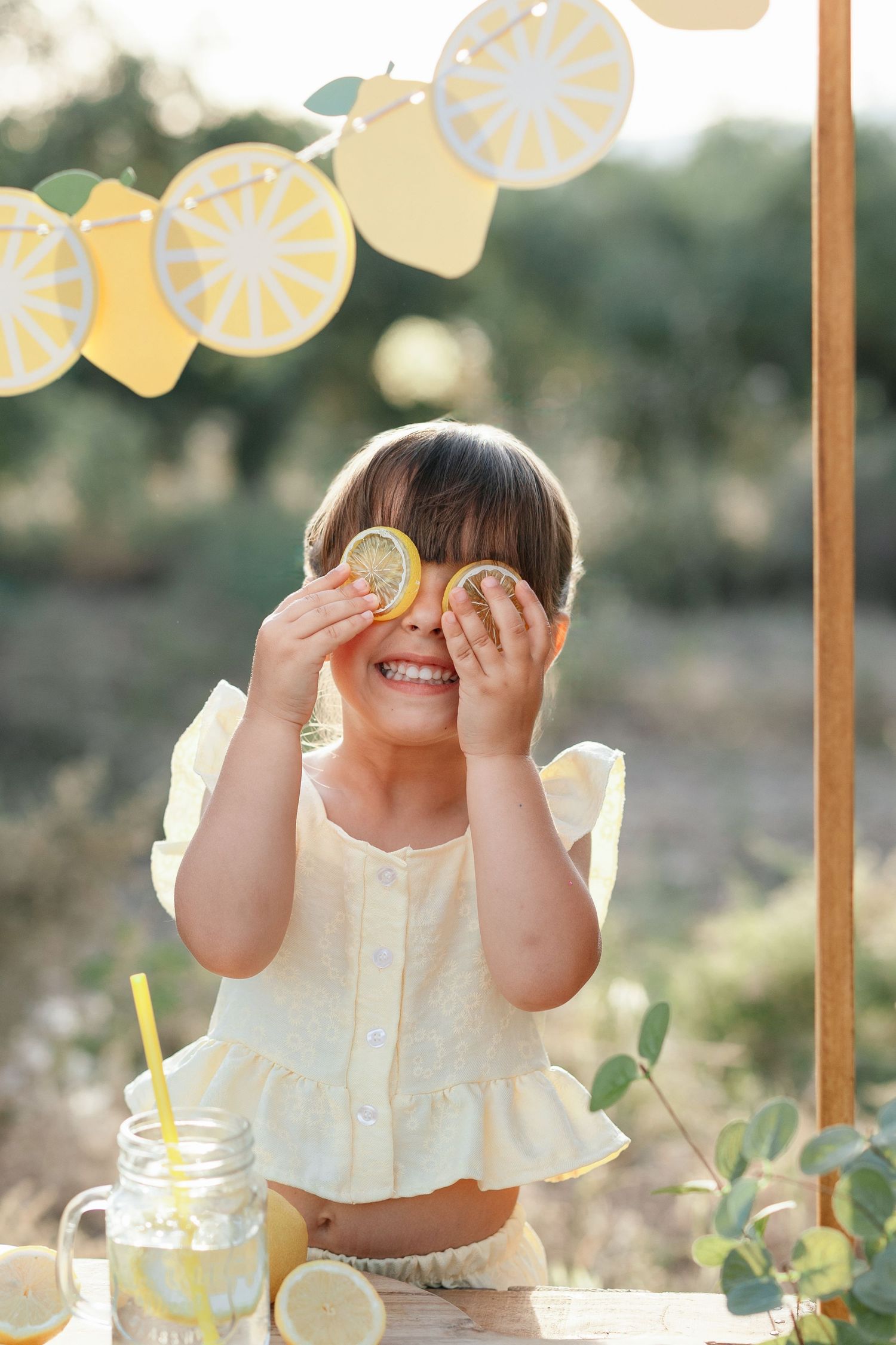 Young girl in yellow top blowing soap bubbles outdoors, surrounded by white balloons and flowers on sunny day.