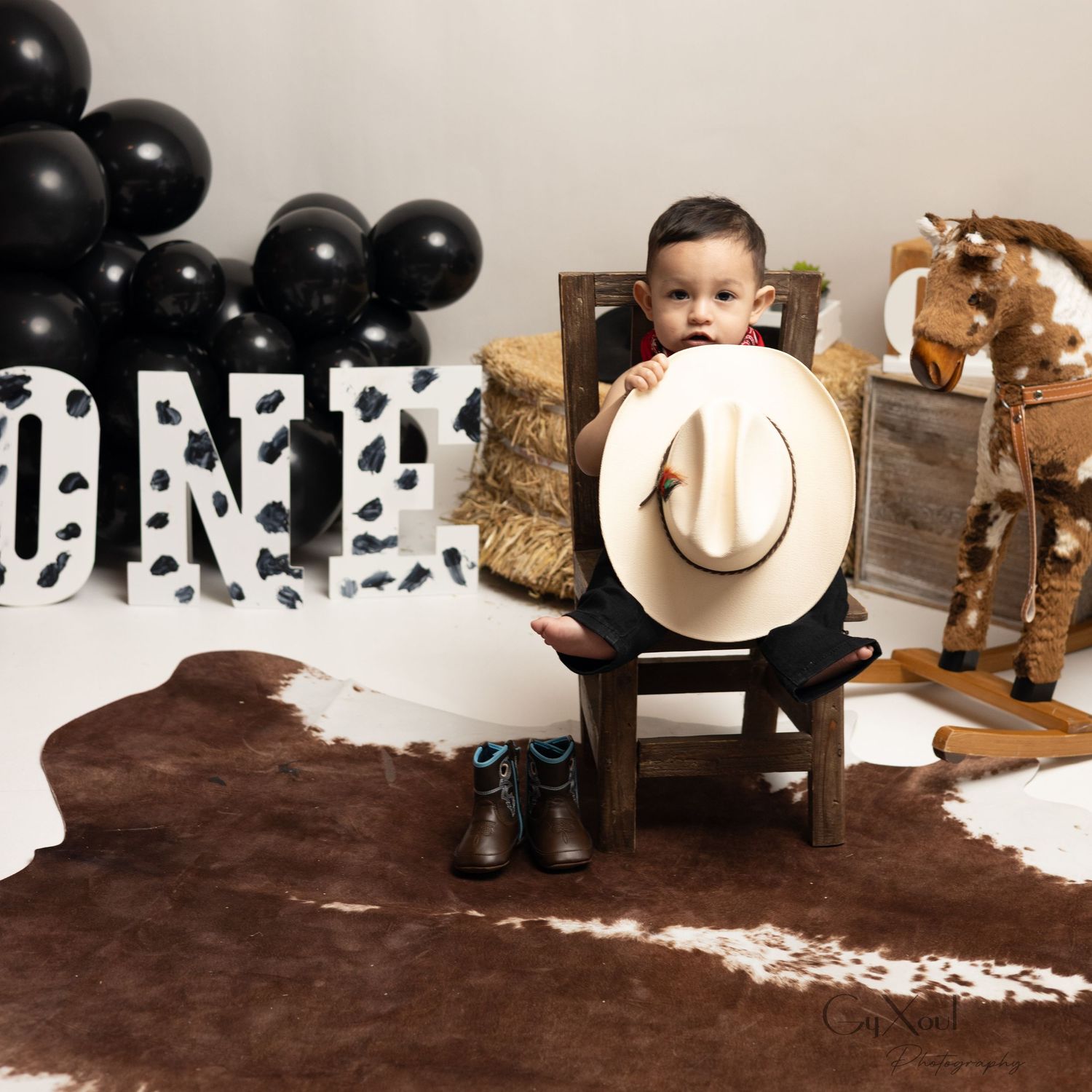 Toddler boy in cowboy boots sits with white hat at western-themed first birthday party with ONE sign and balloons.