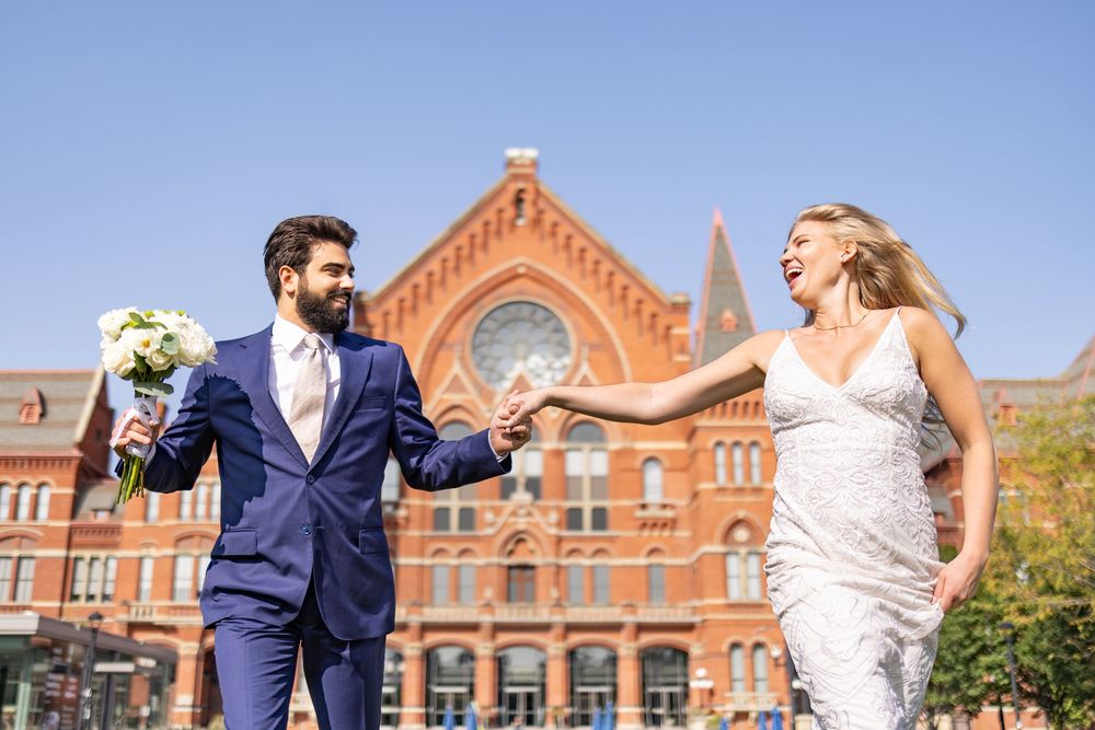 A joyful bride and groom hold hands outside a historic red brick building on their sunny wedding day. Cincinnati Elopement Photographer