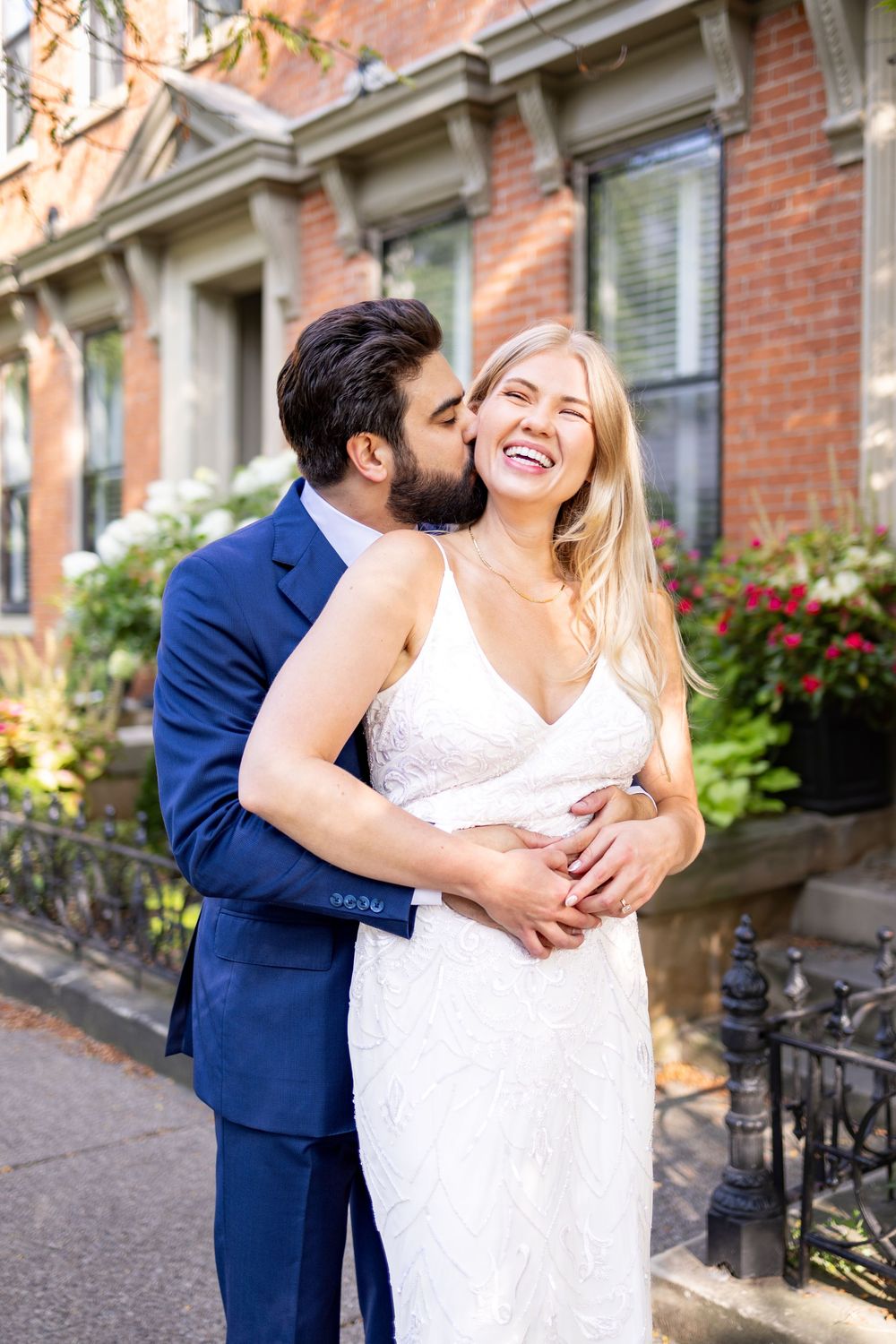 Groom in navy suit kisses bride in white lace gown as she laughs joyfully outside a brick building. Cincinnati Elopement Photographer