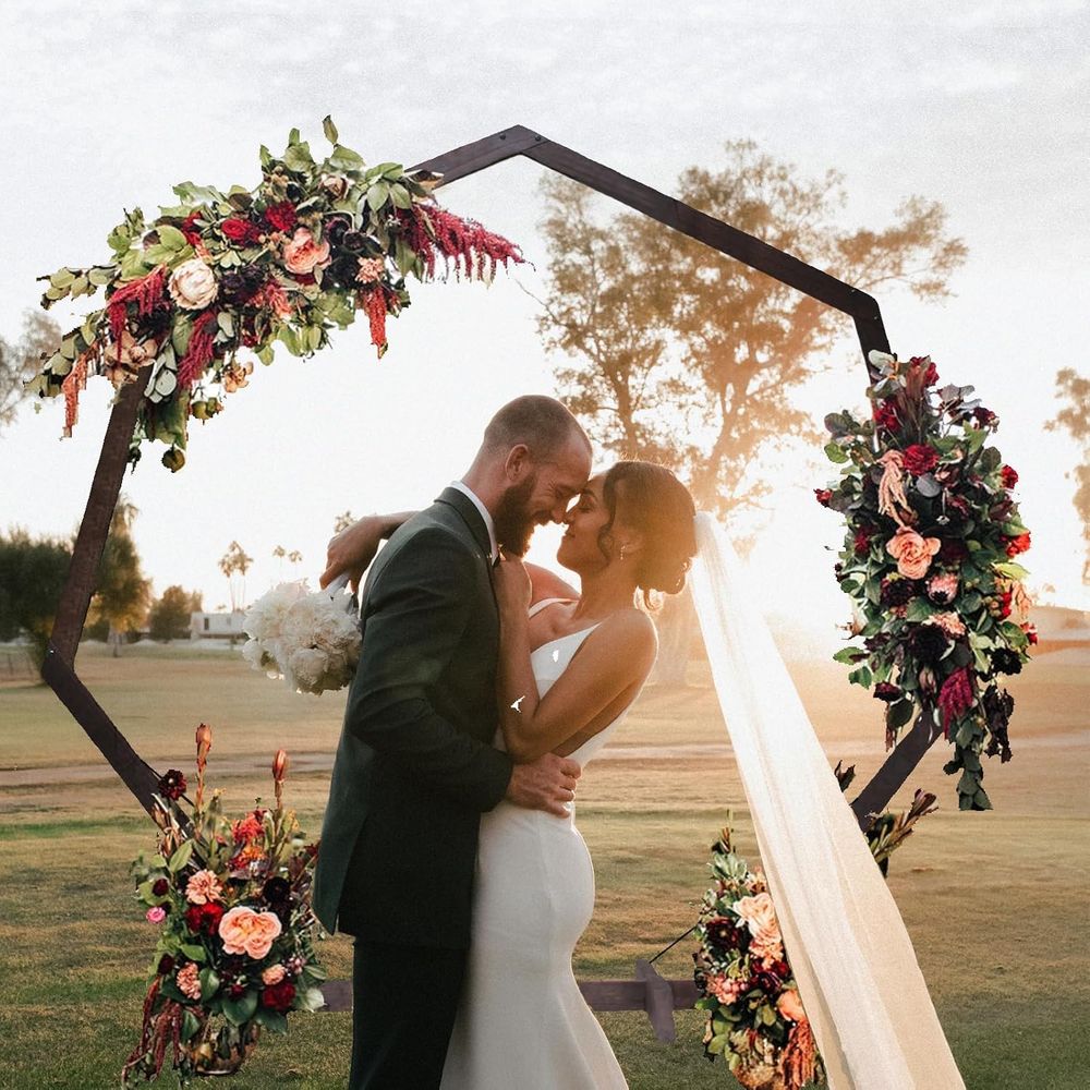 Bride and groom kiss under hexagonal floral arch with burgundy and pink blooms at sunset ceremony.
