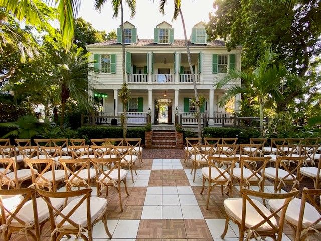 Outdoor wedding ceremony setup with wooden crossback chairs on checkered floor before green historic mansion.