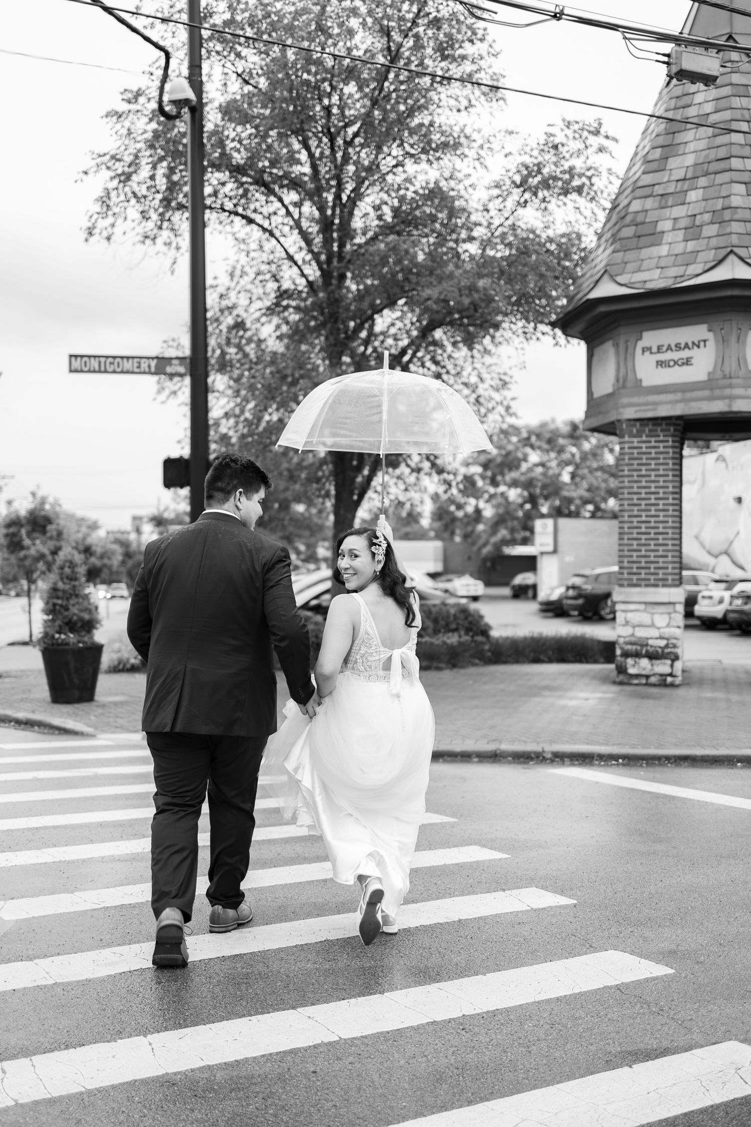 Bride and groom walk hand-in-hand under a clear umbrella across a rainy crosswalk in Pleasant Ridge.
