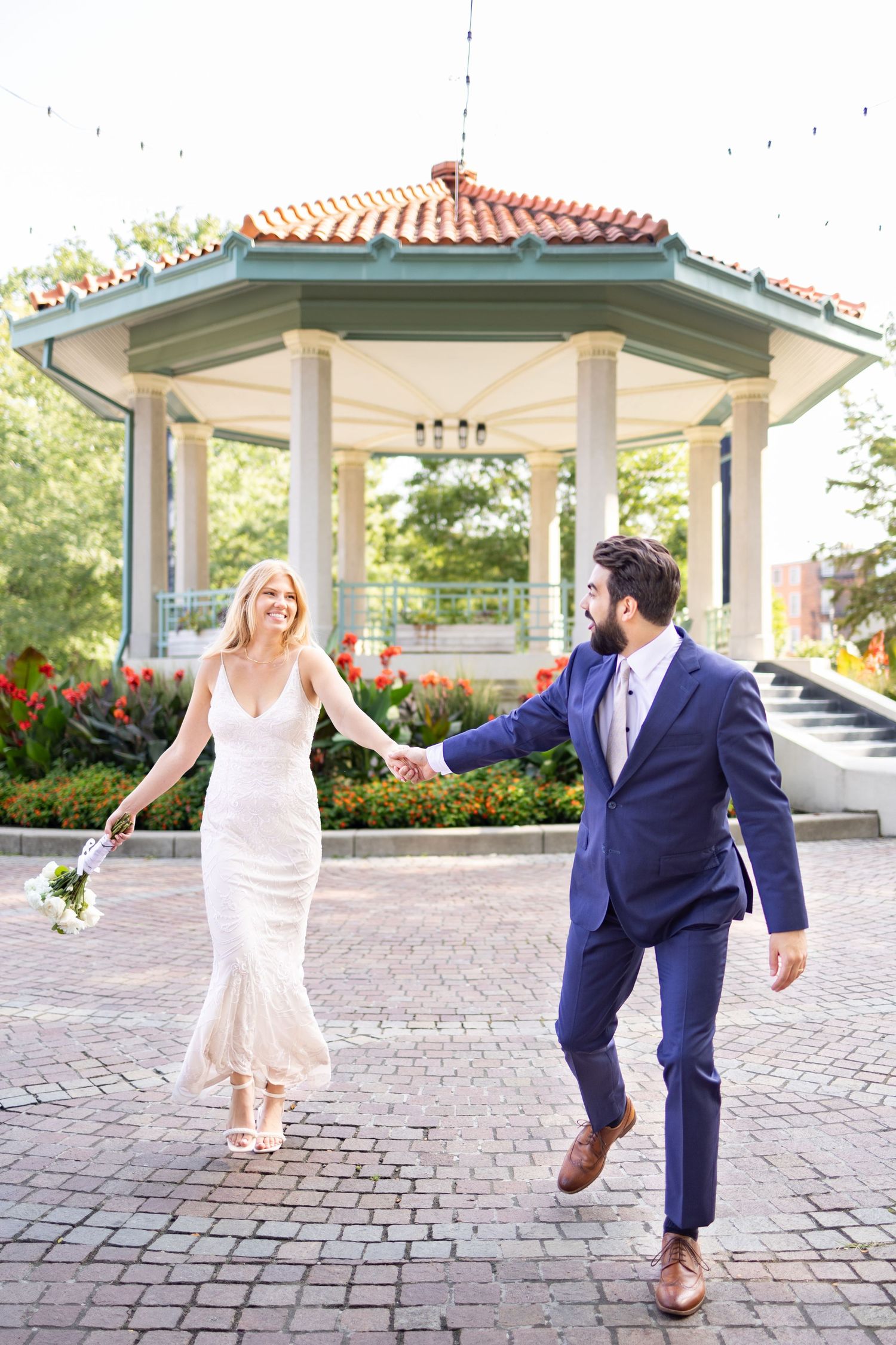 Bride and groom hold hands walking on brick plaza near elegant gazebo at Washington Park Cincinnati
