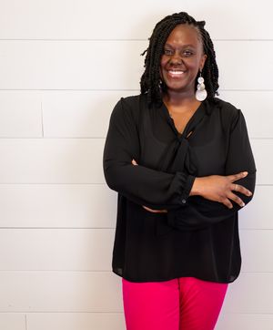 A smiling woman in a black blouse and pink pants stands with arms crossed against a white wall.