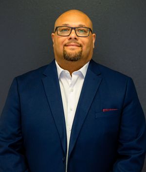 Professional man in navy blue blazer and glasses poses confidently against a gray background for a business portrait.