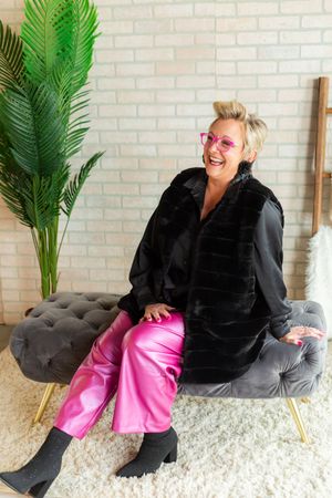 Woman laughing in pink sunglasses, shiny pink pants, black fur vest, seated on gray tufted bench near tropical plant.