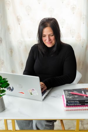 Woman in black turtleneck typing on sticker-covered laptop at white desk with books nearby.