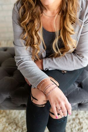 Woman wearing layered rose gold bracelets and rings, seated in gray jeans and striped cardigan.