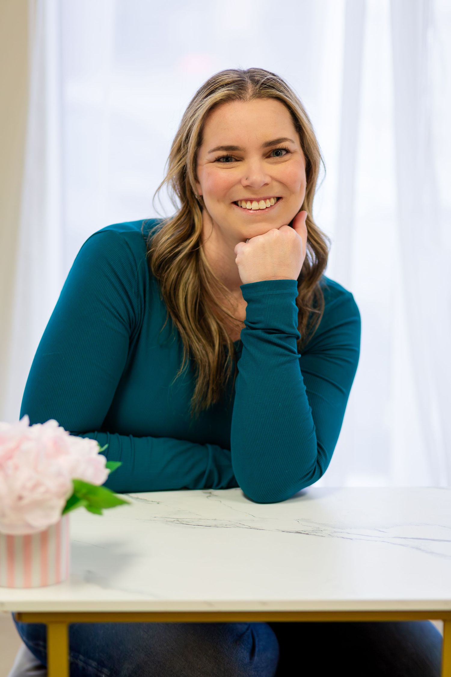 Smiling woman in teal top resting chin on hand at white table with pink flowers, bright natural light background.