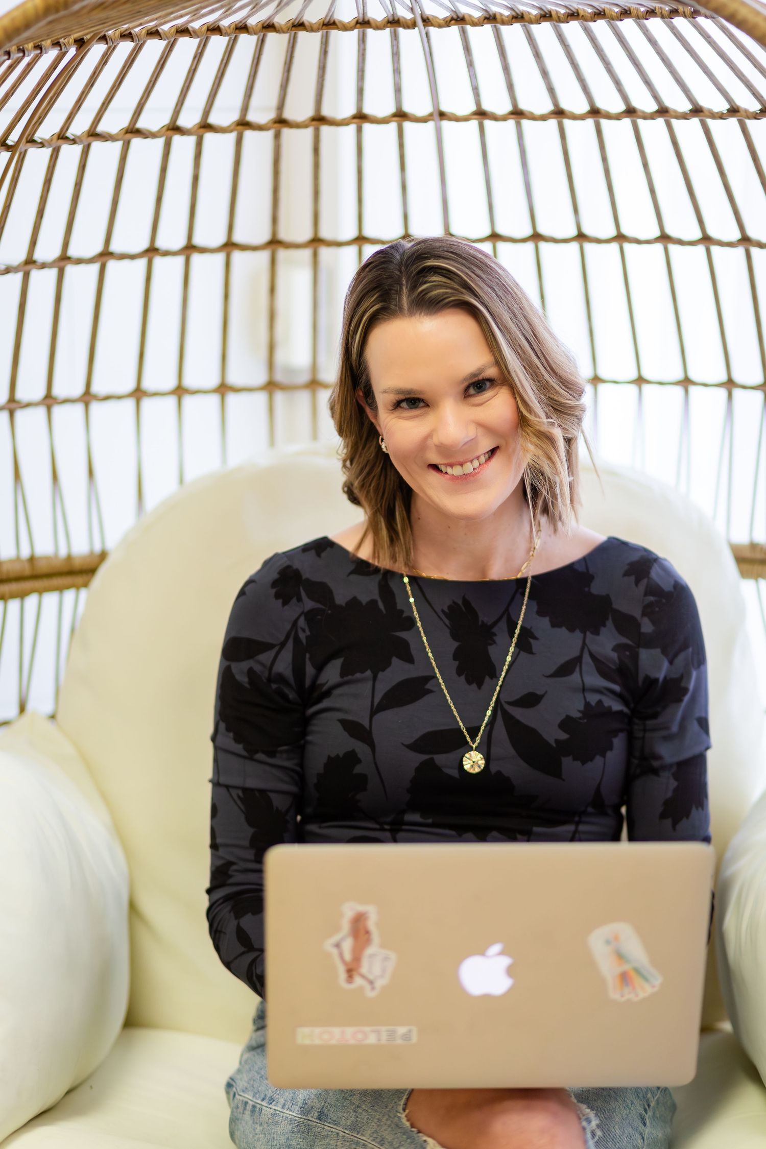 Smiling woman with laptop sitting in a rattan egg chair wearing a black floral top and gold necklace.