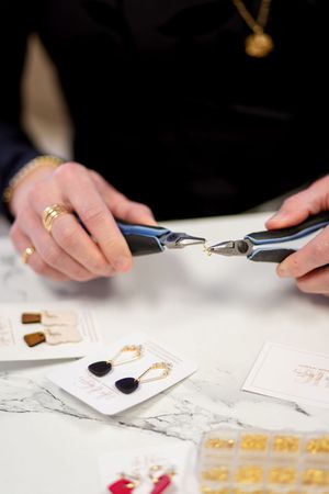 Jeweler using pliers to assemble gold drop earrings with dark stones on a marble surface.