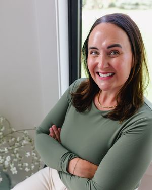 Smiling woman with dark hair in green long-sleeve top, arms crossed, posing near a window in a bright room.