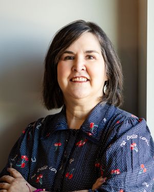 Smiling woman with dark hair wearing a navy floral blouse, arms crossed, posed against a neutral background.