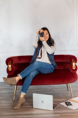 Woman sitting on red velvet sofa holding camera with MacBook laptop on floor in elegant white room.