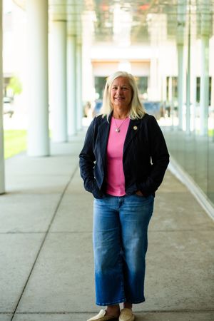 Smiling blonde woman in pink top, navy blazer, and wide-leg jeans standing in modern building corridor.