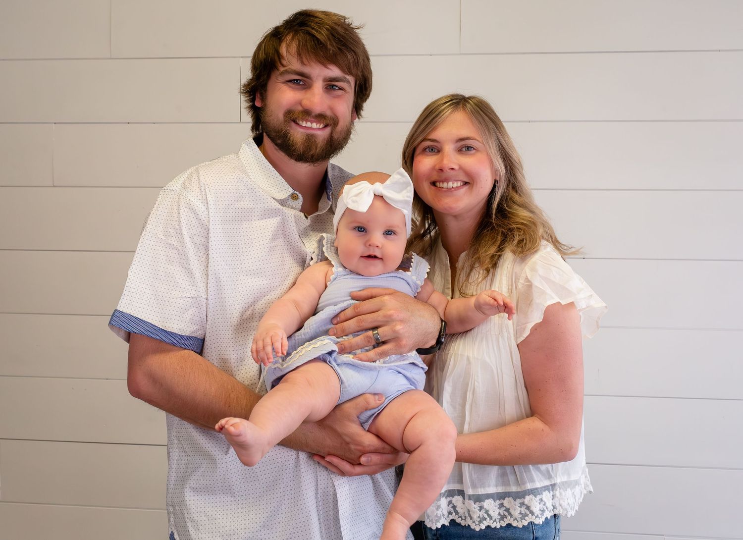 A smiling young family with a man, woman, and baby girl in a bow posing together against a white shiplap wall.