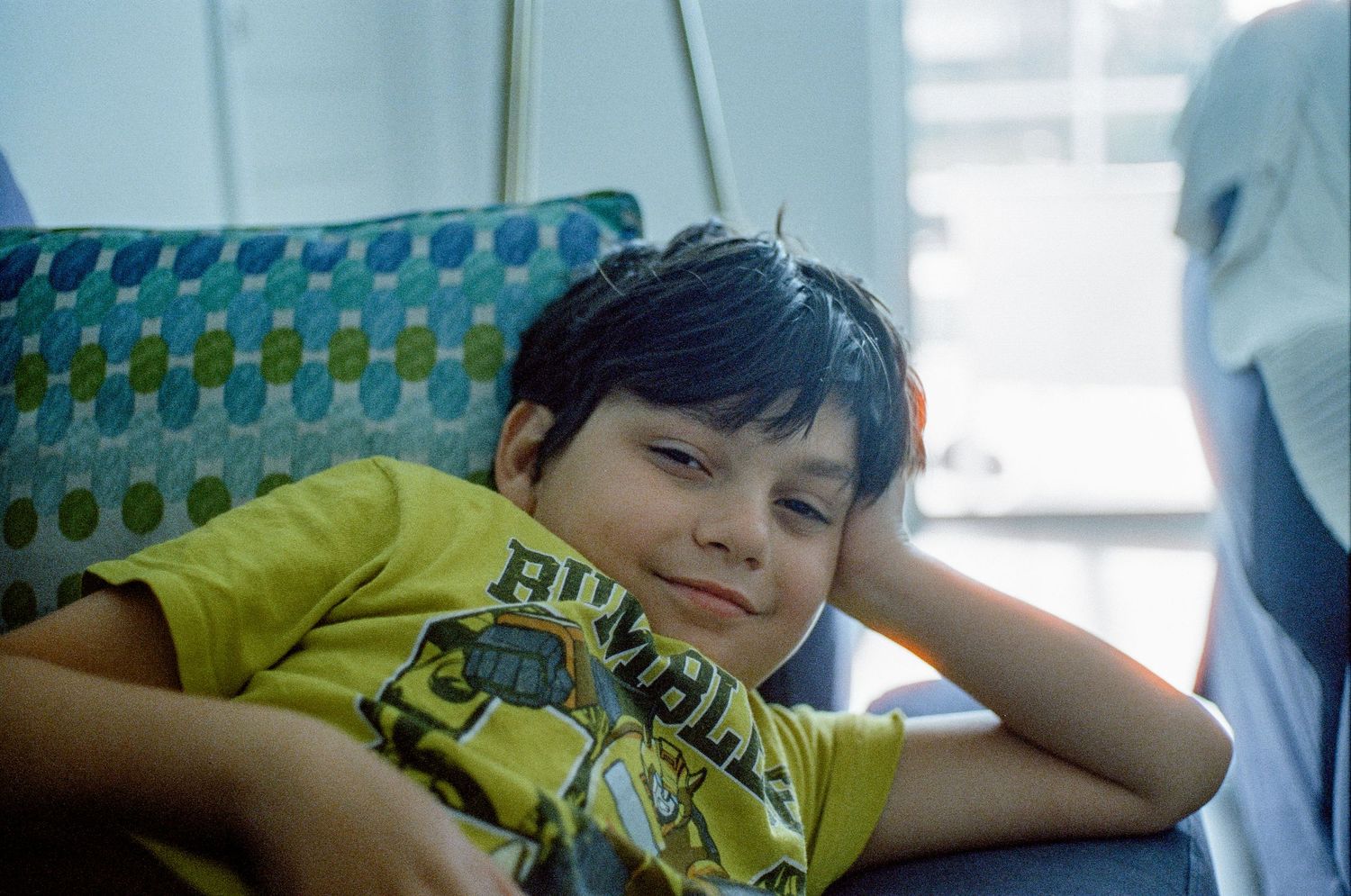 A young child in a yellow t-shirt rests their head on their arm while sitting near a window.