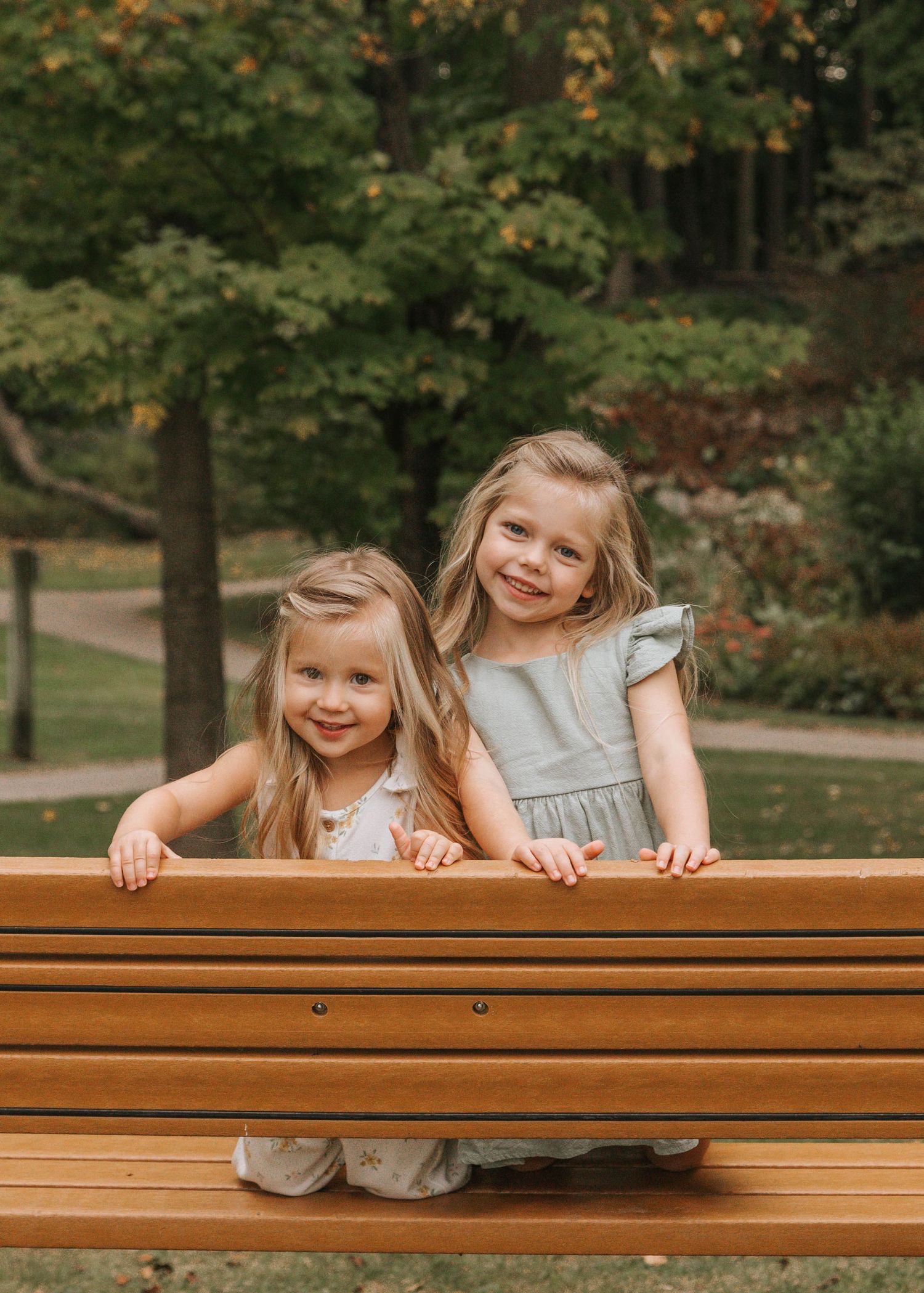 Playful children peeking over wooden park bench surrounded by autumn trees.