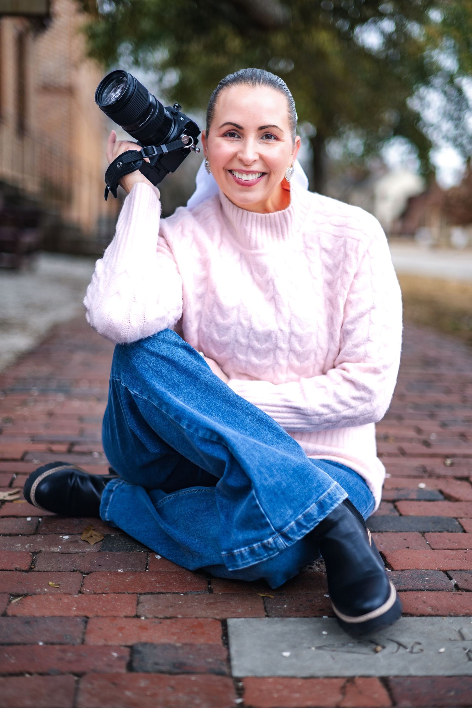 Photographer sitting cross-legged on brick walkway wearing pink cable knit sweater, jeans and holding professional camera.