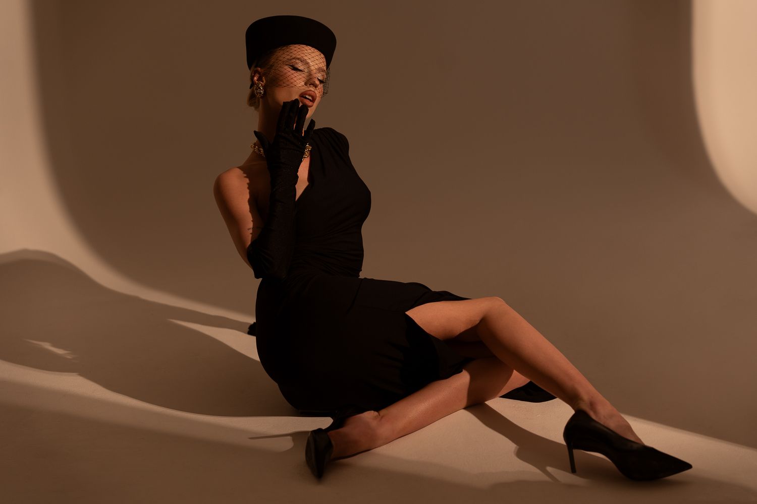 A person in vintage fashion wearing a black pillbox hat with veil, black gloves and dress against a warm sepia background.