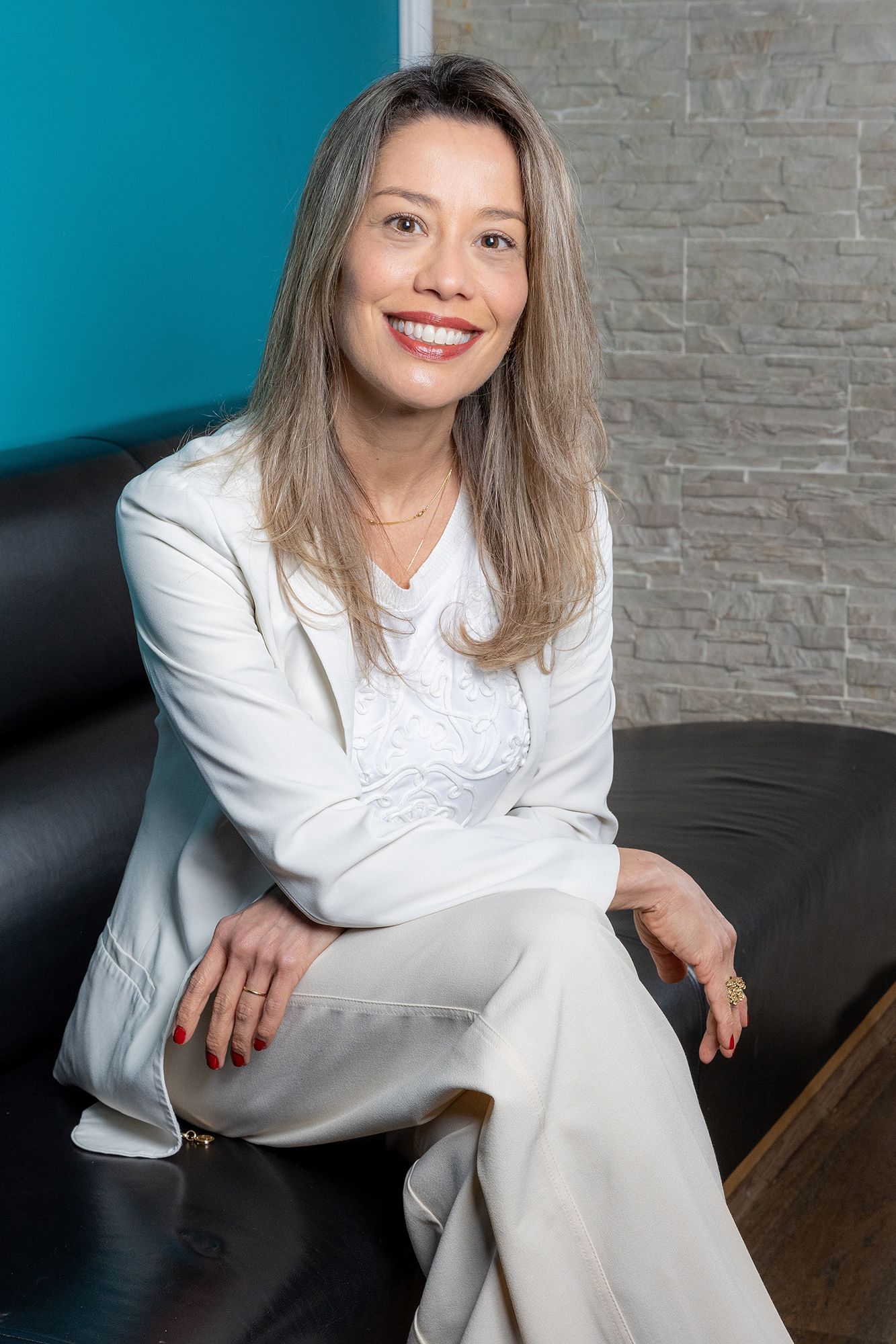 Professional business portrait of someone wearing white sitting relaxed on a dark couch against a teal and gray wall.