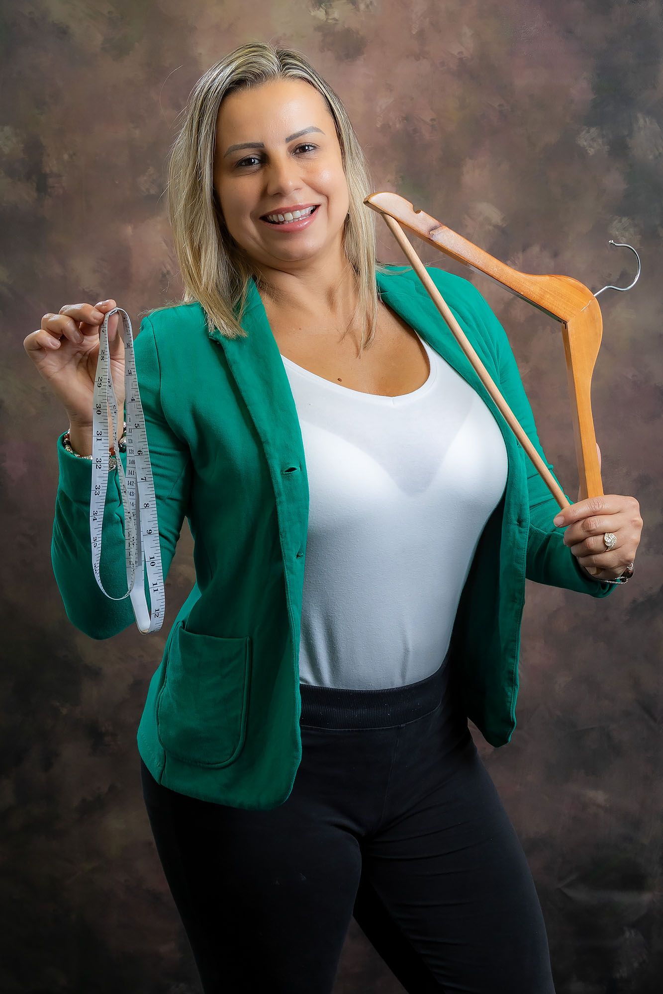 A person wearing a bright green blazer and white top holds up clothing hangers against a dark studio backdrop.