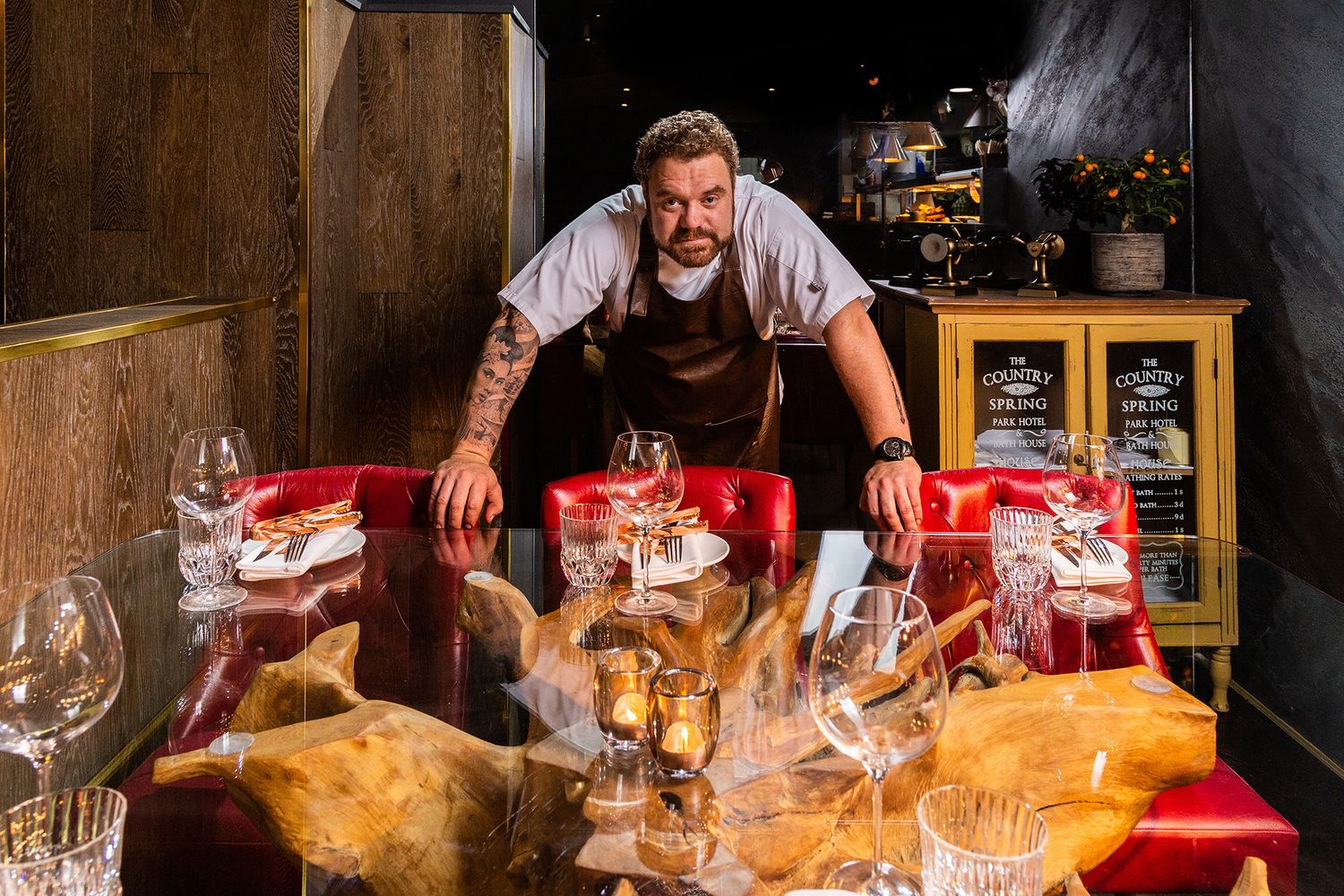 Bartender stands behind table with wine glasses and cheese plates in rustic wooden setting with dramatic lighting.