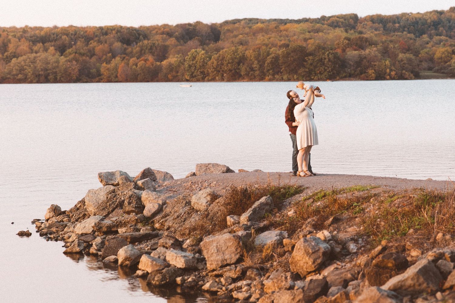 A family standing on a rocky pier near a lake. The man has his arms around the woman who is holding a baby up in the air like an airplane.