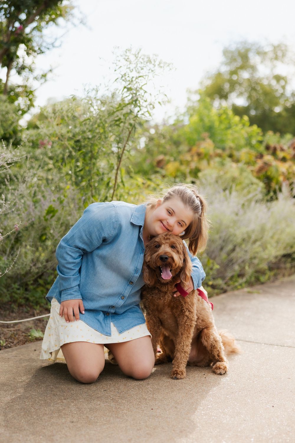 Child in blue shirt sitting with a brown fluffy dog on a pathway surrounded by greenery.