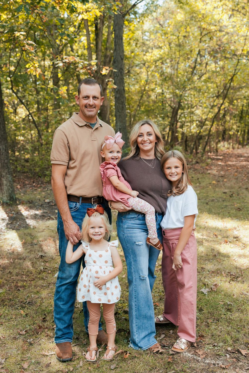 Family group portrait in casual attire standing together in a sunlit wooded area.