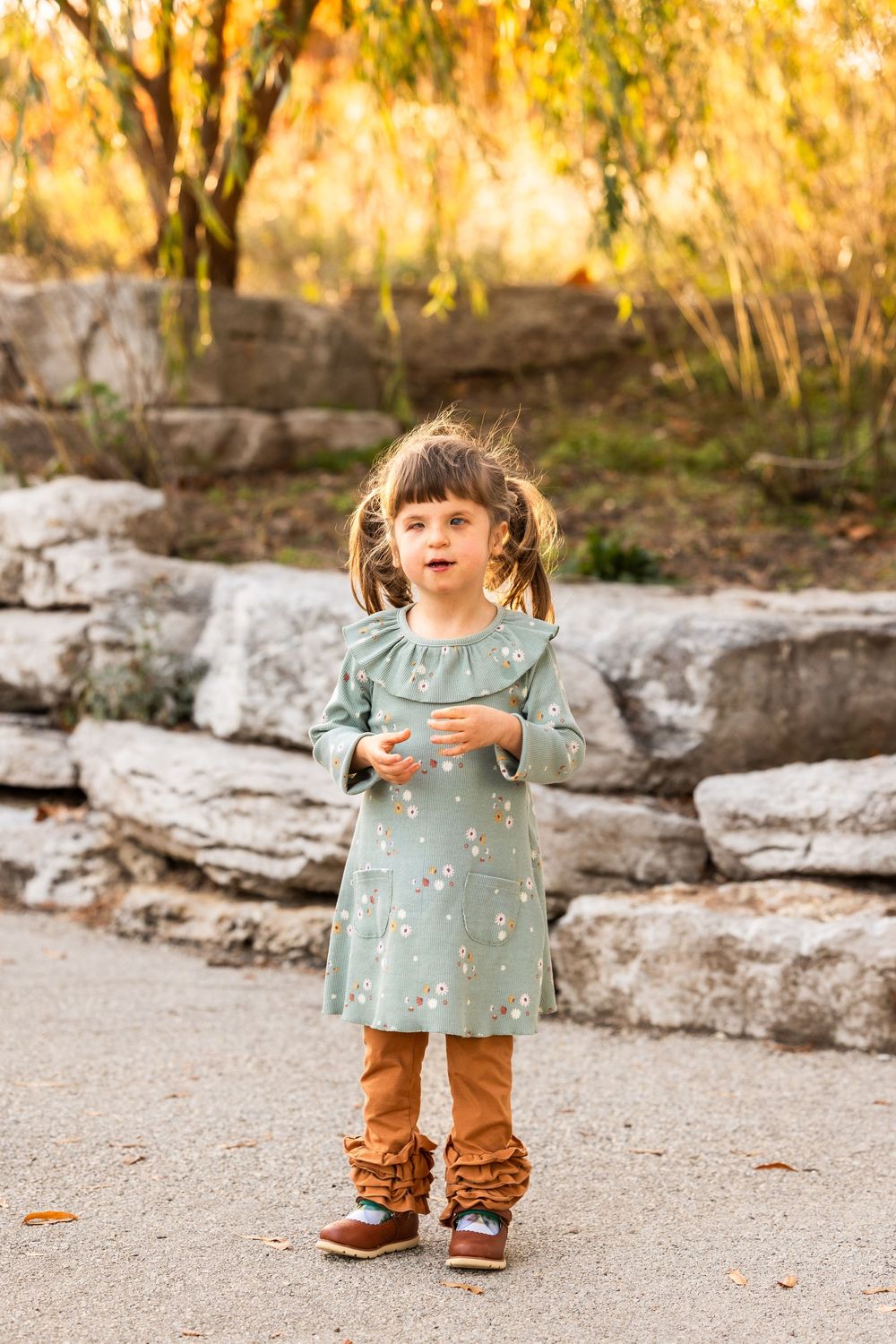 Toddler in teal dress and brown pants walking on stone path outdoors.