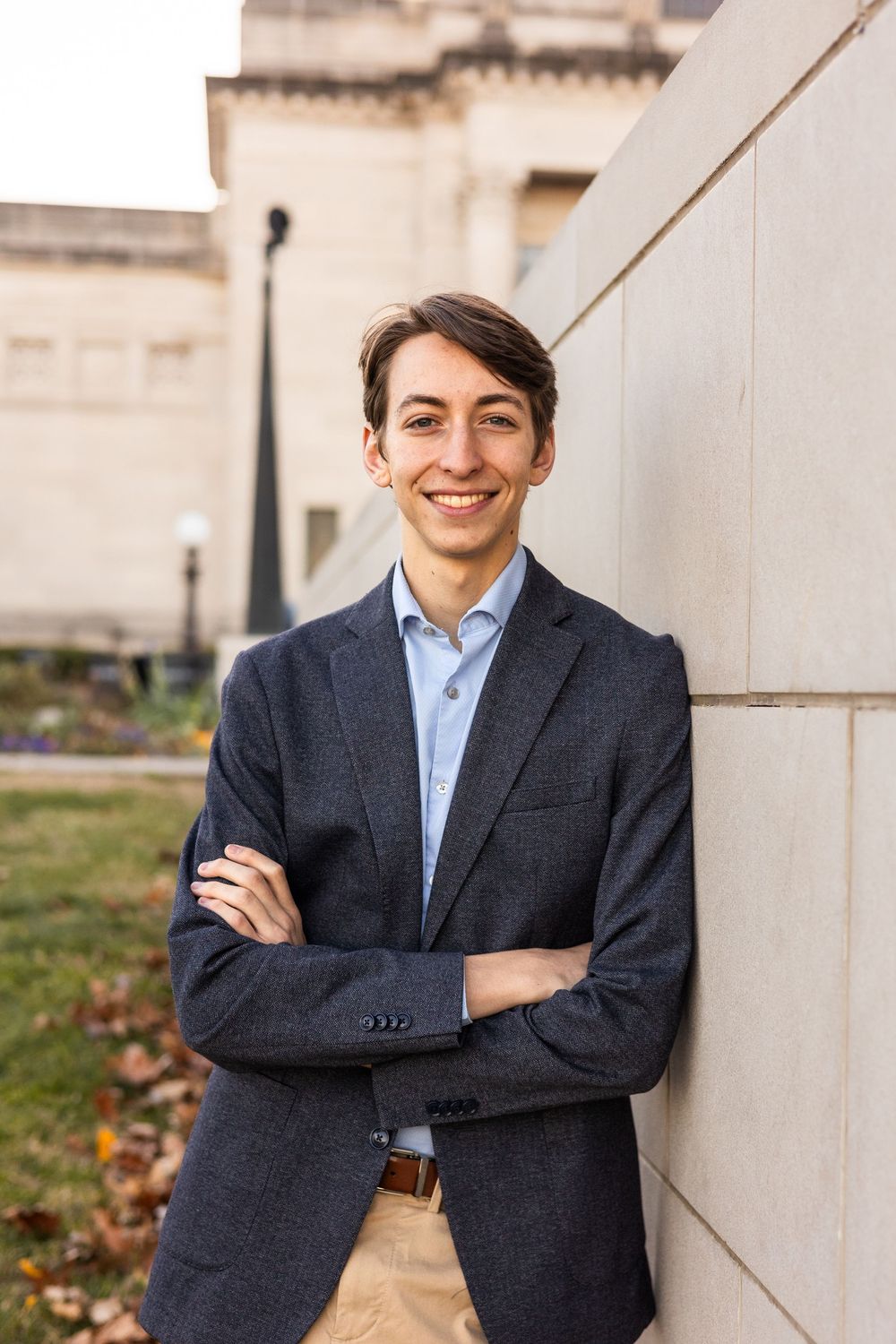 Professional portrait of someone in a navy blazer and blue shirt leaning against a wall.
